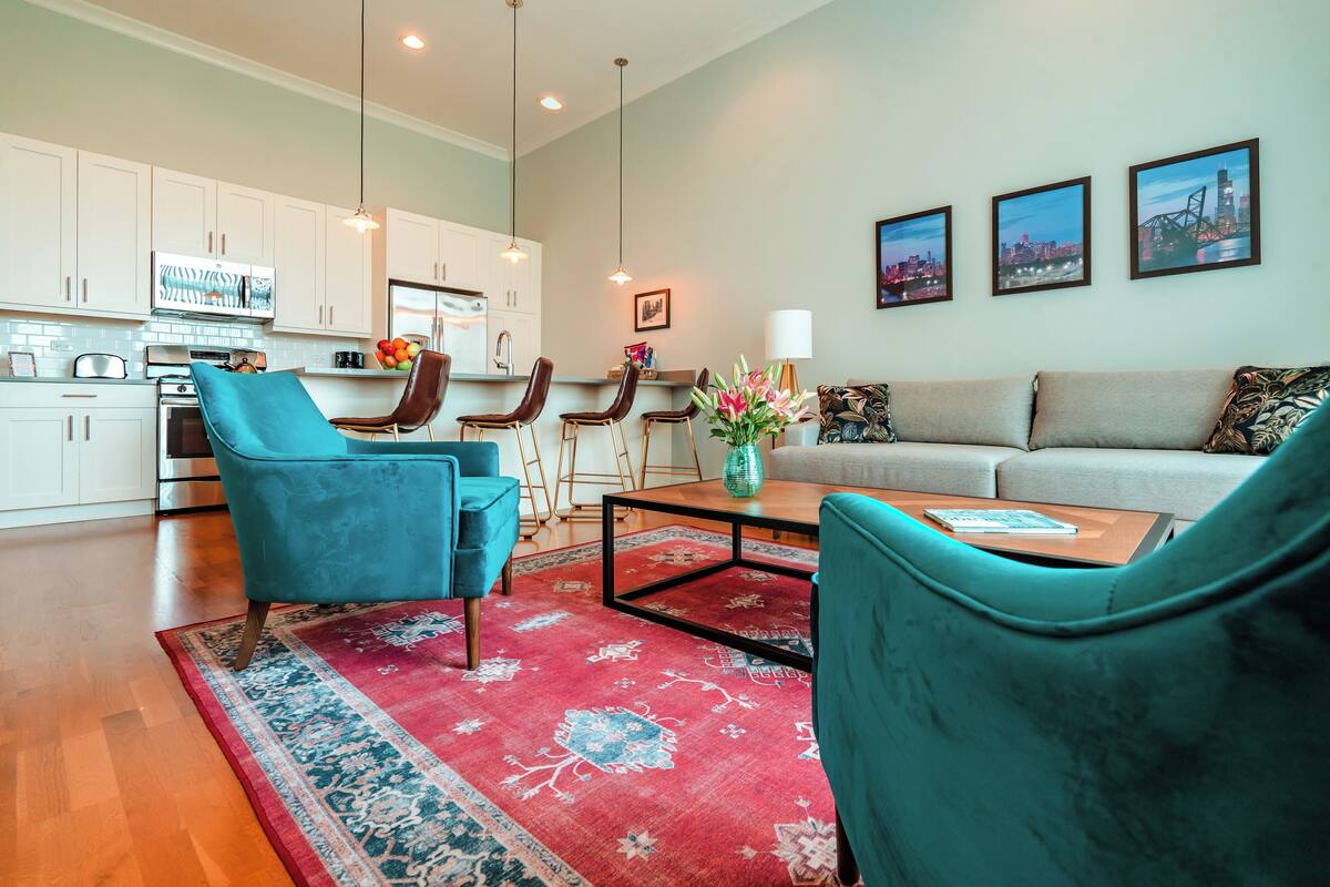 A modern living room with a sofa, two chairs, a coffee table, and a rug. There is a kitchen area with white cabinets and a dining table with chairs.