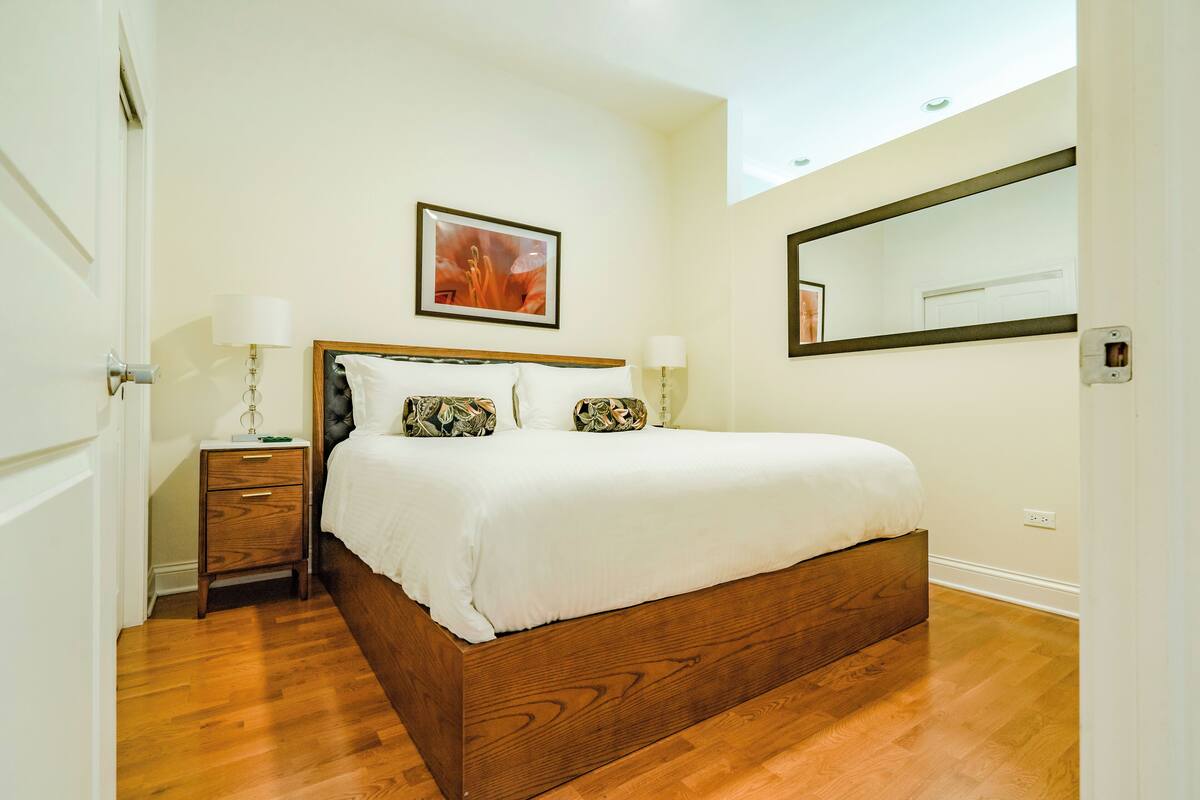 A bedroom with a wooden bed, two bedside tables with lamps, a framed picture on the wall, and a large mirror.
