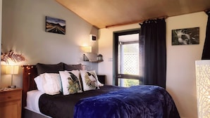A bedroom with a bed, a nightstand, a lamp, a window with curtains, and wall art. - Height of Dunedin (North Taieri)