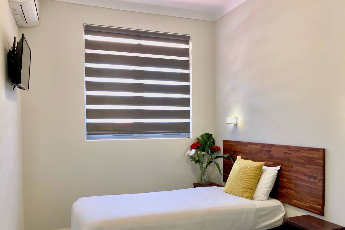 A neatly arranged hotel room with a single bed, a bedside table, a nightstand, a wall-mounted TV, a window with blinds, and a vase with flowers.