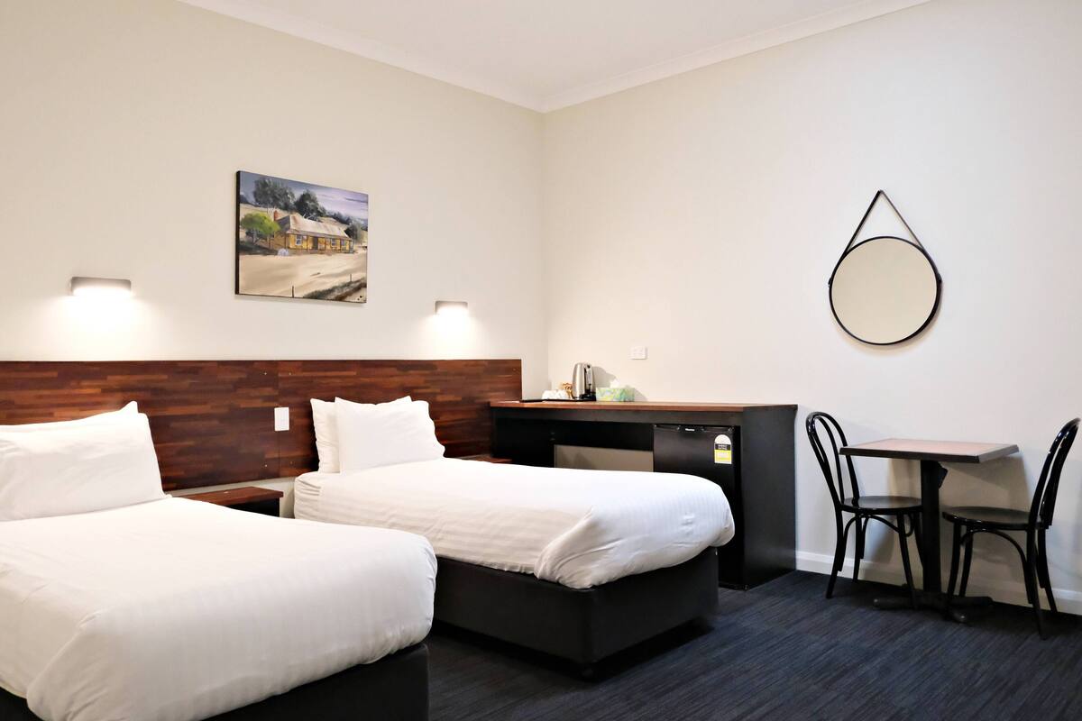 A hotel room with two beds, a desk, a chair, a small table, a mirror, and a painting on the wall.