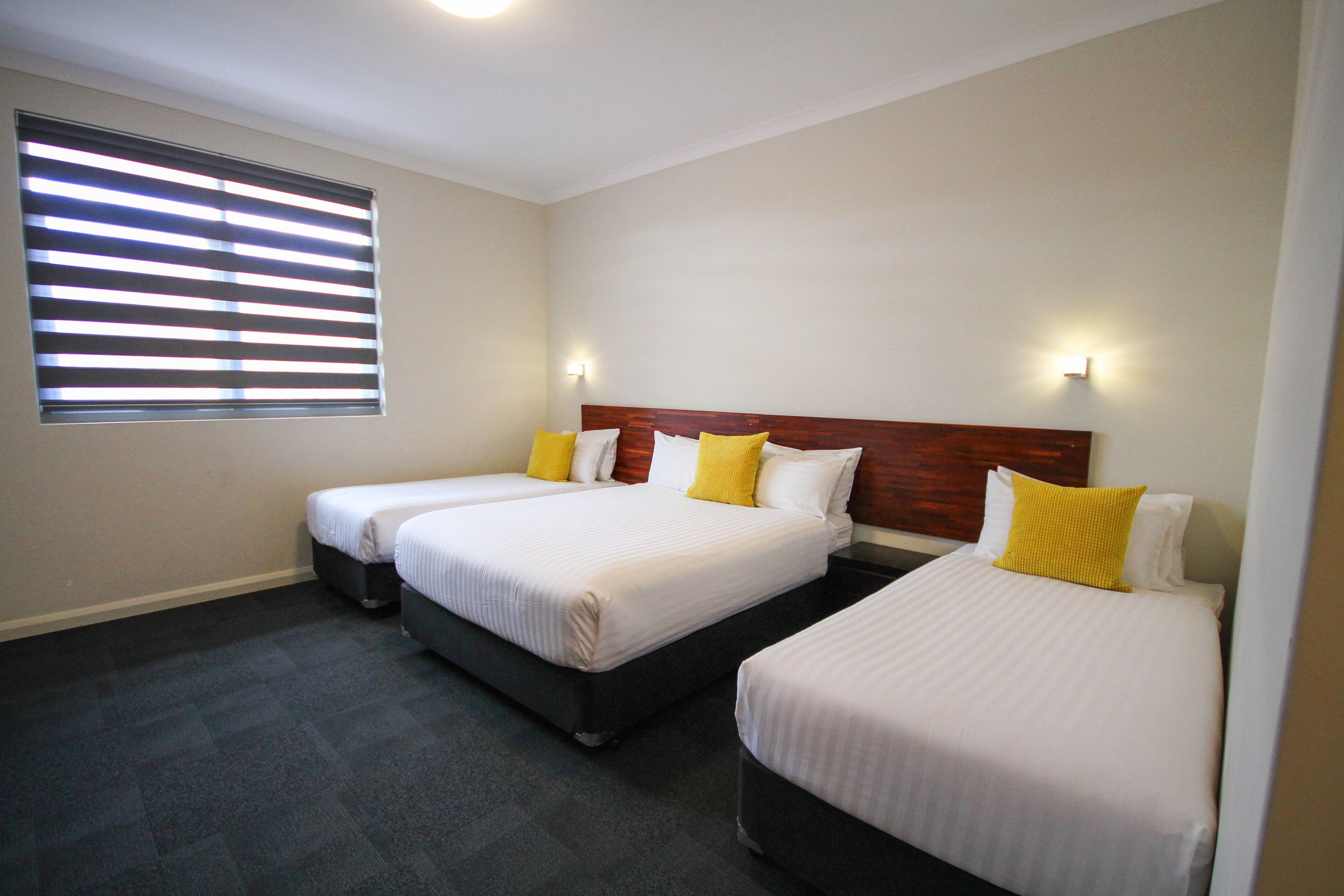 Habitación de hotel con dos camas, cada una con ropa blanca y almohadas amarillas, un ventanal grande con persianas y un suelo alfombrado oscuro.