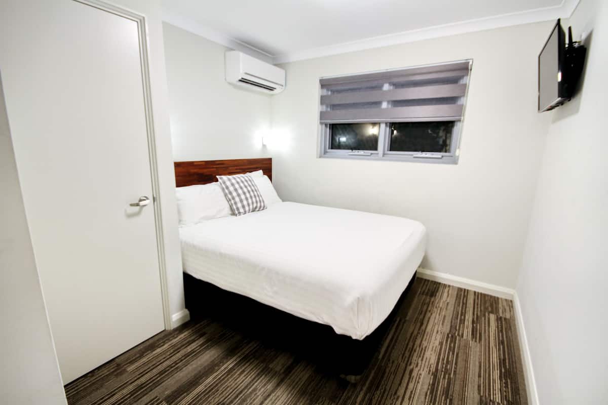A small, neatly arranged bedroom with a single bed, a window with blinds, an air conditioning unit, and a wall-mounted TV.