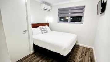 Une petite chambre à coucher, bien rangée, avec un lit simple, une fenêtre avec des stores, un climatiseur et un téléviseur fixé au mur.