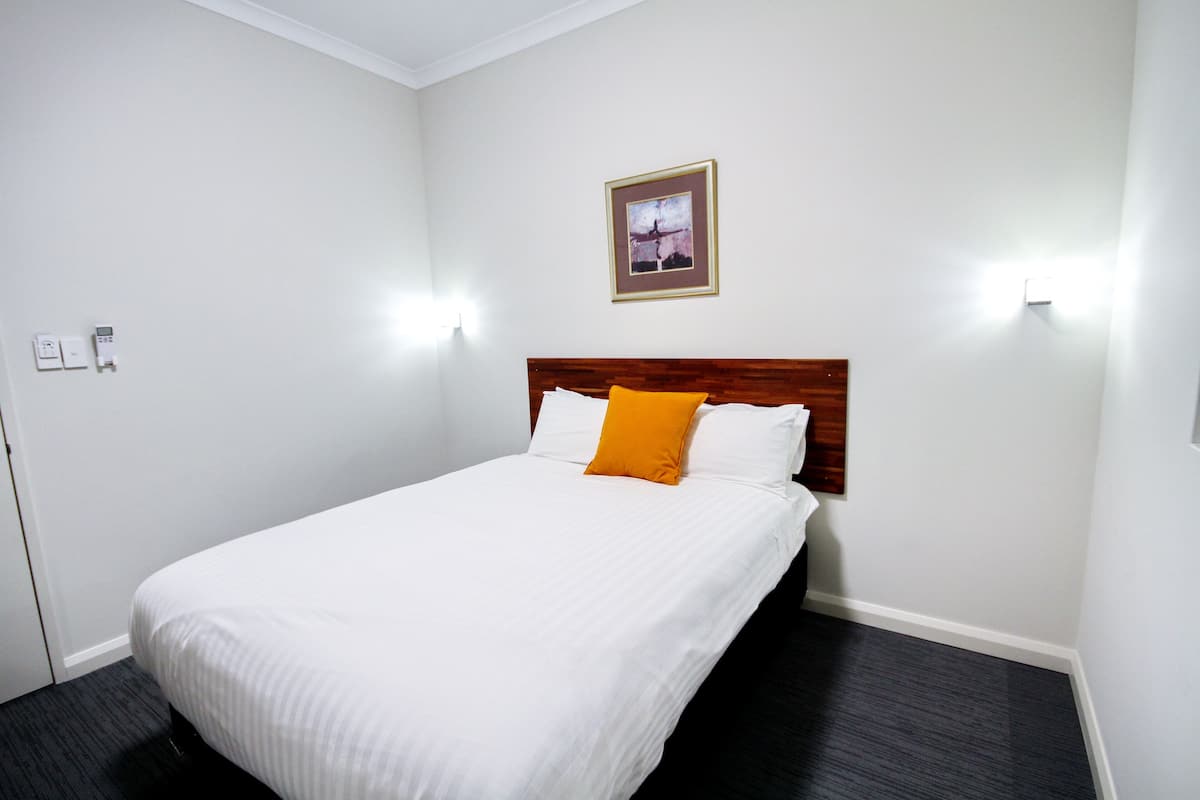 A hotel room with a single bed, white bedding, a wooden headboard, an orange accent pillow, and a framed picture on the wall.