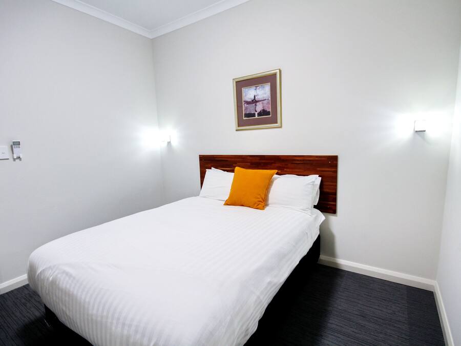 Une chambre d’hôtel avec un lit simple, du linge de lit blanc, une tête de lit en bois, un coussin décoratif orange et un tableau encadré au mur.
