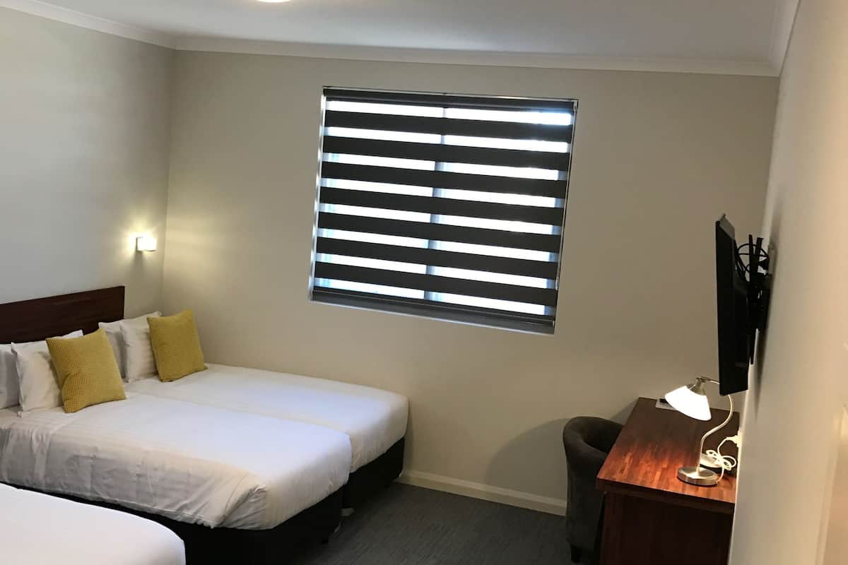 A hotel room with two single beds, a desk with a lamp, a window with blinds, and a ceiling light.