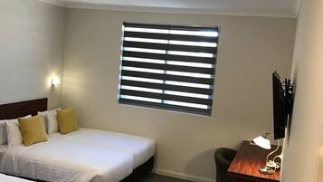 A hotel room with two single beds, a desk with a lamp, a window with blinds, and a ceiling light.