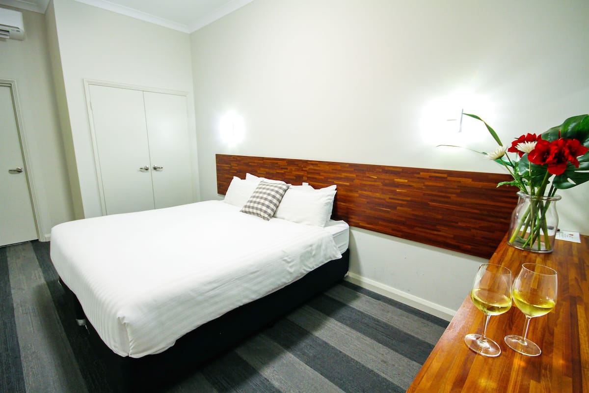 A hotel room with a bed, a wooden headboard, a nightstand with two wine glasses, and a vase with flowers.