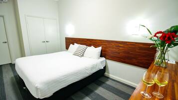 A hotel room with a bed, a wooden headboard, a nightstand with two wine glasses, and a vase with flowers.