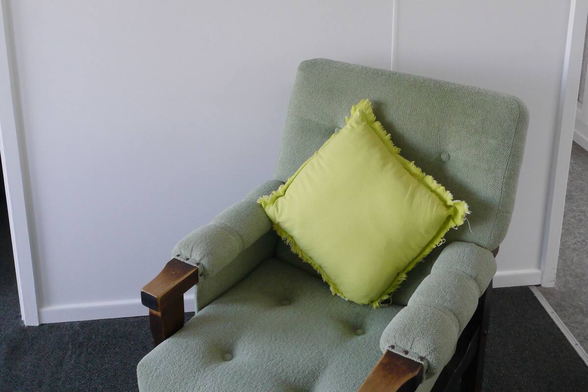 Un sillón verde claro con un cojín amarillo frente a una pared blanca, con un cuadro de una escena de playa en la parte superior.