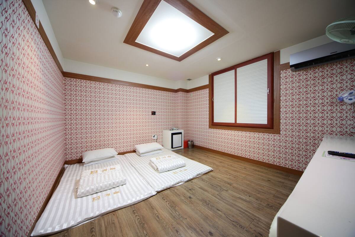A room with a skylight, patterned wallpaper, a bed with pillows, a small bedside table, and a window with blinds.