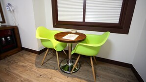 A small room with a round wooden table and two bright green chairs. A window with blinds is on the wall, and a hairdryer is mounted on the wall to the left. - Castle Motel Iksan (Iksan)