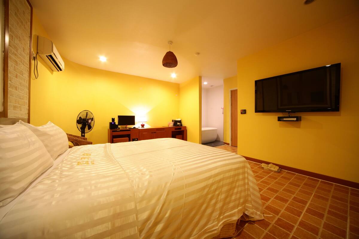 A bedroom with a bed, a desk, a chair, a television, and an air conditioning unit.