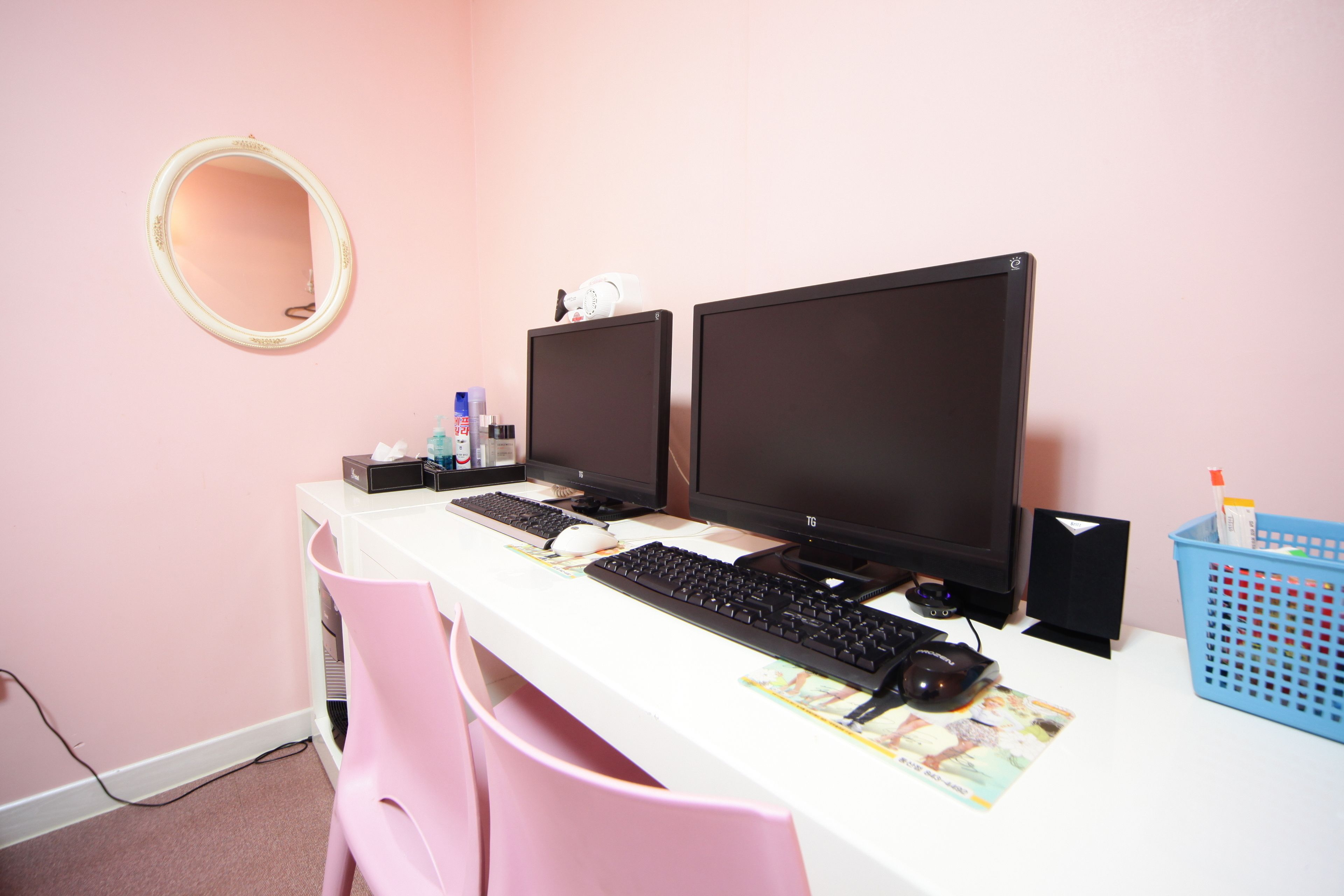 Una habitación con un escritorio blanco, dos monitores, un teclado, un ratón, un espejo y una silla rosa.