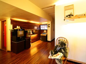 A hotel lobby with a wooden reception desk, a coffee machine, a television, and a teddy bear reading a magazine.