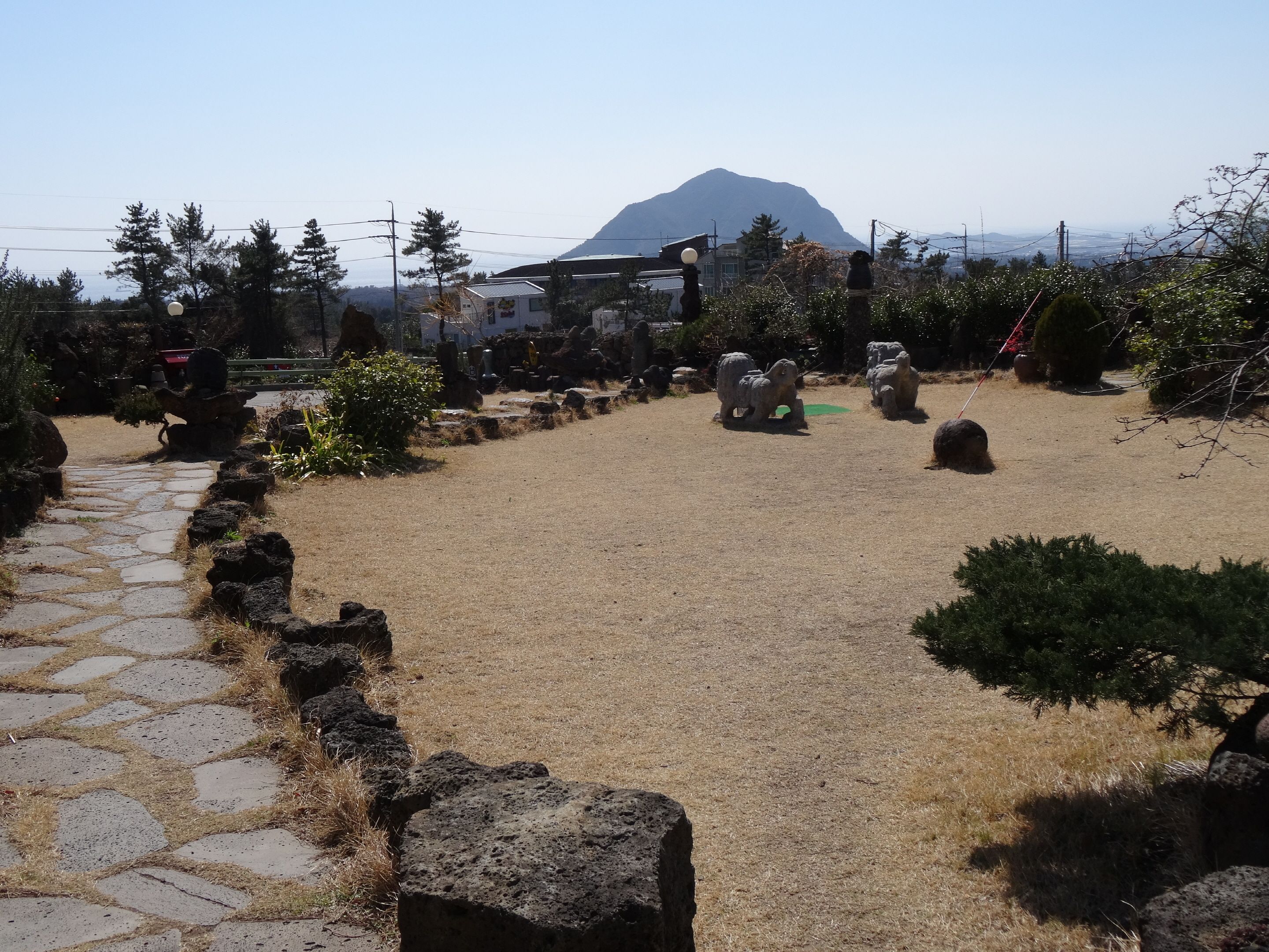 A stone pathway leading to a grassy area with sculptures and a mountain in the background.