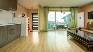A modern kitchen with wooden cabinets and a built-in oven, a dining area with a glass table, and a living room with a sofa and a view of the outside. - Lionhill Pension (Seogwipo)