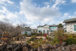 A residential area with stone houses, a paved driveway, and a garden with various plants and rocks. - Lionhill Pension (Seogwipo)