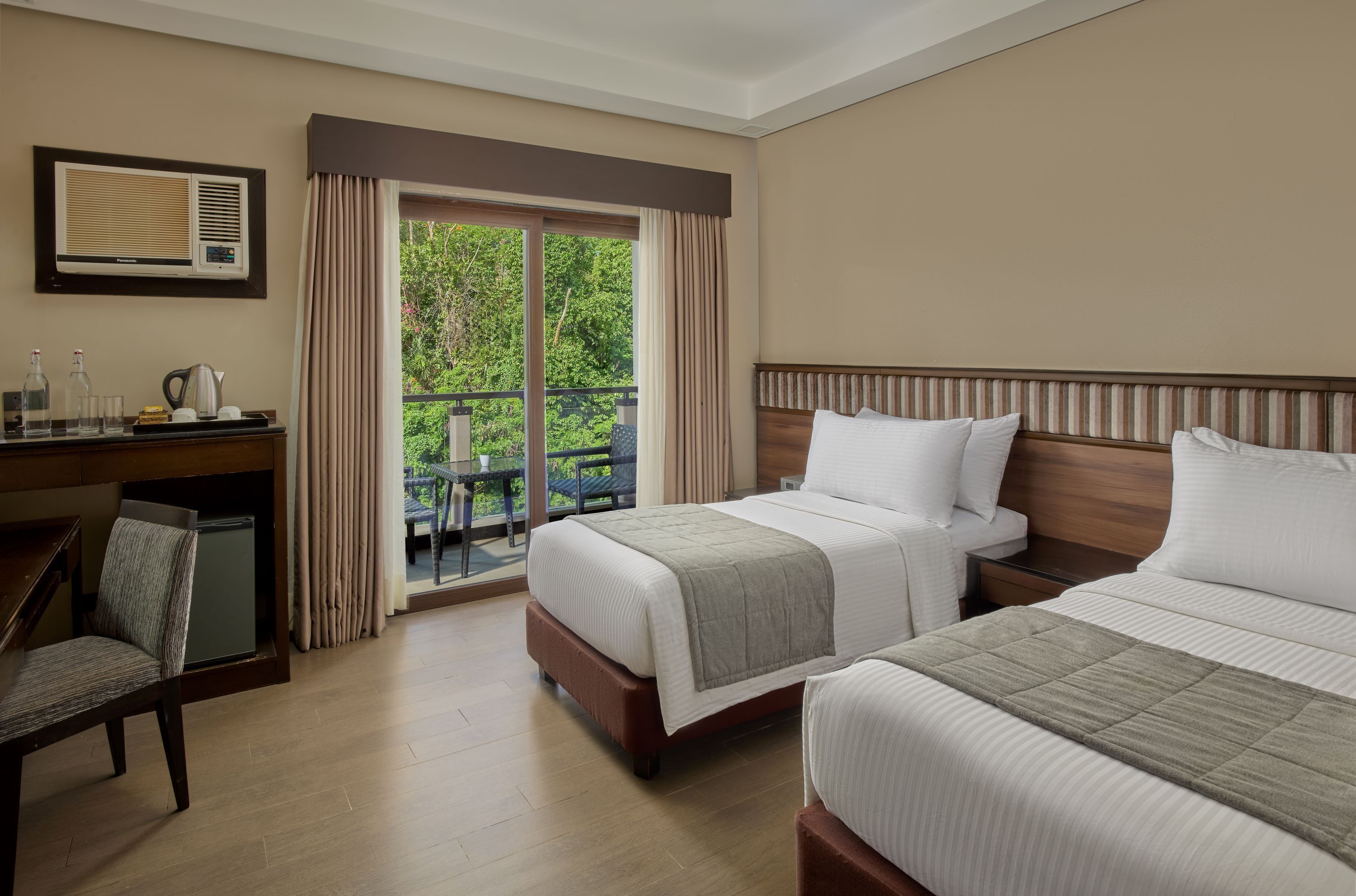 Habitación de hotel con dos camas, un escritorio, una silla, ventana con cortinas y un balcón con vistas a la vegetación.