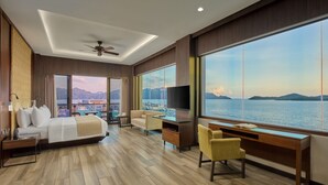 Una habitación de hotel moderna con cama, escritorio, televisor y un amplio ventanal con vistas al mar y las montañas.