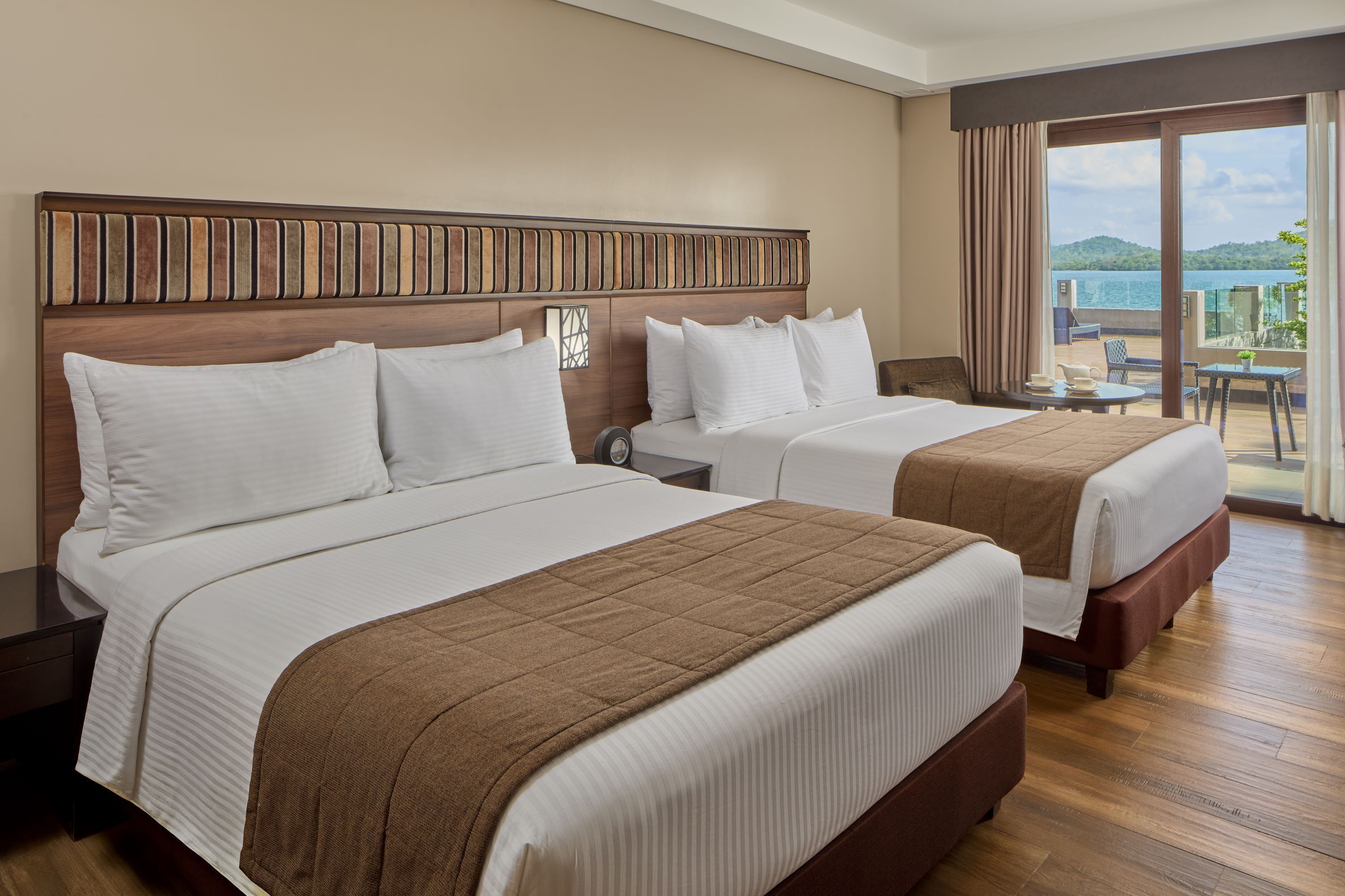 Ein Hotelzimmer mit zwei Betten, einem Holzkopfteil, einem kleinen Tisch mit Stühlen und einem Balkon mit Meerblick.