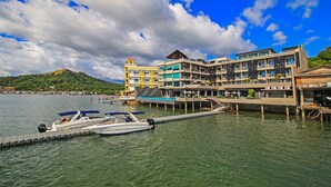 Un hotel frente al agua con varios pisos, un muelle con un bote, y una montaña al fondo.