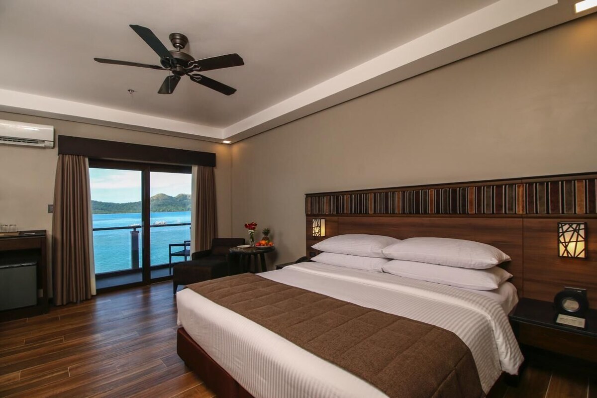 A hotel room with a bed, a ceiling fan, a nightstand, a desk, and a view of the sea.