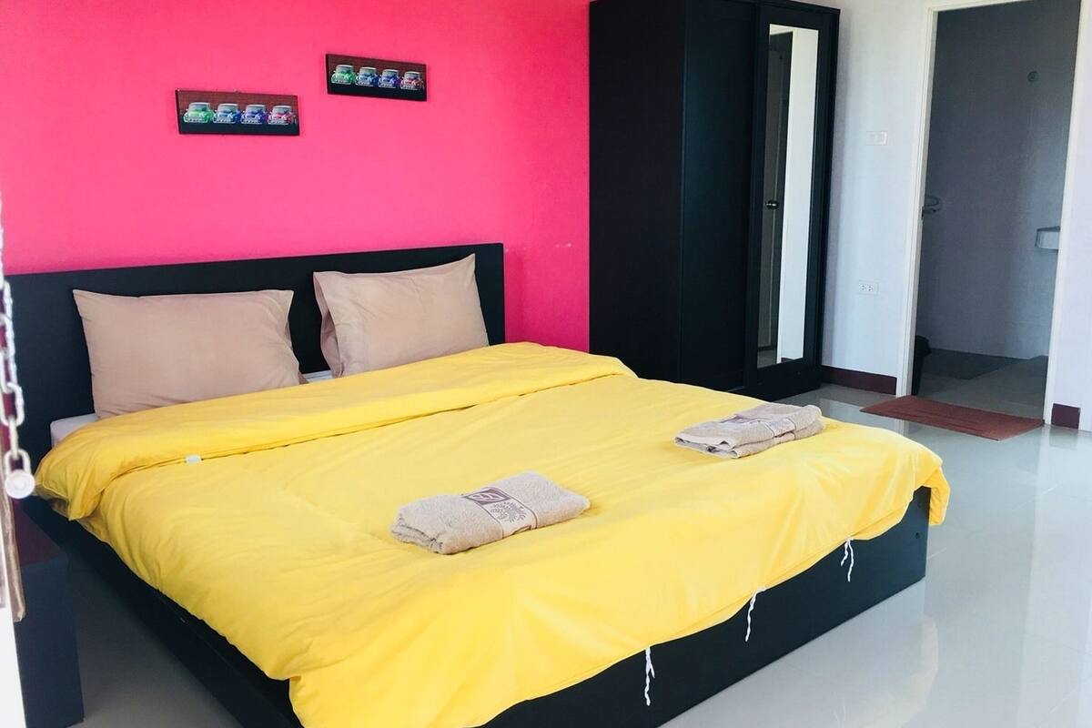 A bed with a yellow bedspread and two pillows, a black wardrobe, and a bright pink wall with car stickers.