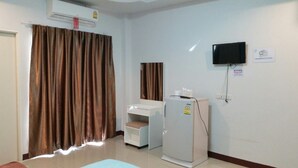 A room with a large window covered by brown curtains, a white desk with a chair, a white cabinet, a white refrigerator, and a mounted flat-screen TV. - The Rich Home (Nakhon Ratchasima)