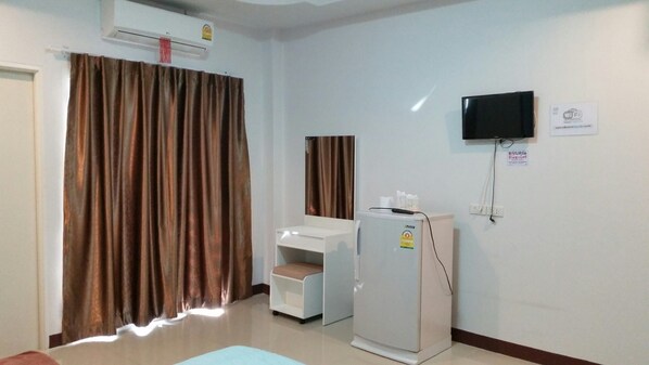 A room with a large window covered by brown curtains, a white desk with a chair, a white cabinet, a white refrigerator, and a mounted flat-screen TV.