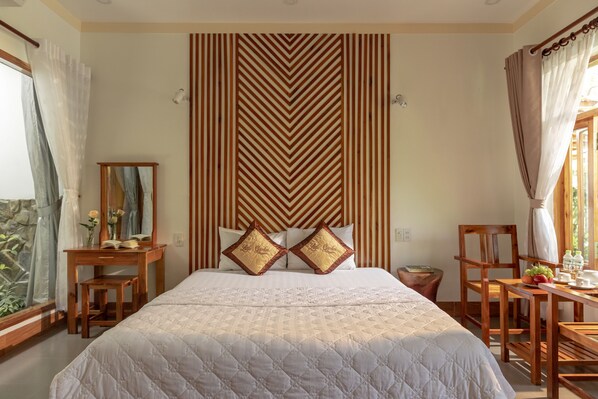 A hotel room with a large bed, a wooden desk, a chair, and a table with fruits and a glass. - Little Garden Bungalow (Phu Quoc)