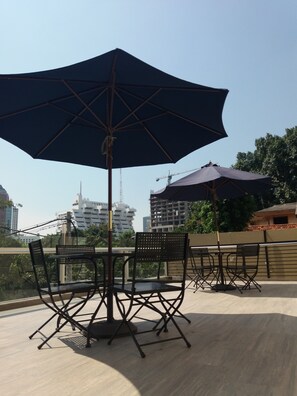 An outdoor seating area with tables and chairs under large umbrellas, overlooking a cityscape with buildings and a bridge. - Moscha Hotel Surabaya (Surabaya)