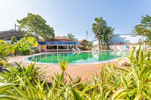 A swimming pool with a tiled deck, surrounded by lush greenery and a building with a blue roof.
