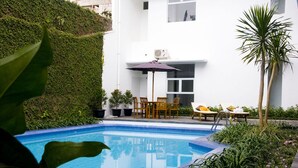 Una piscina con solárium de baldosas, rodeada de follaje y un edificio blanco de techo plano.