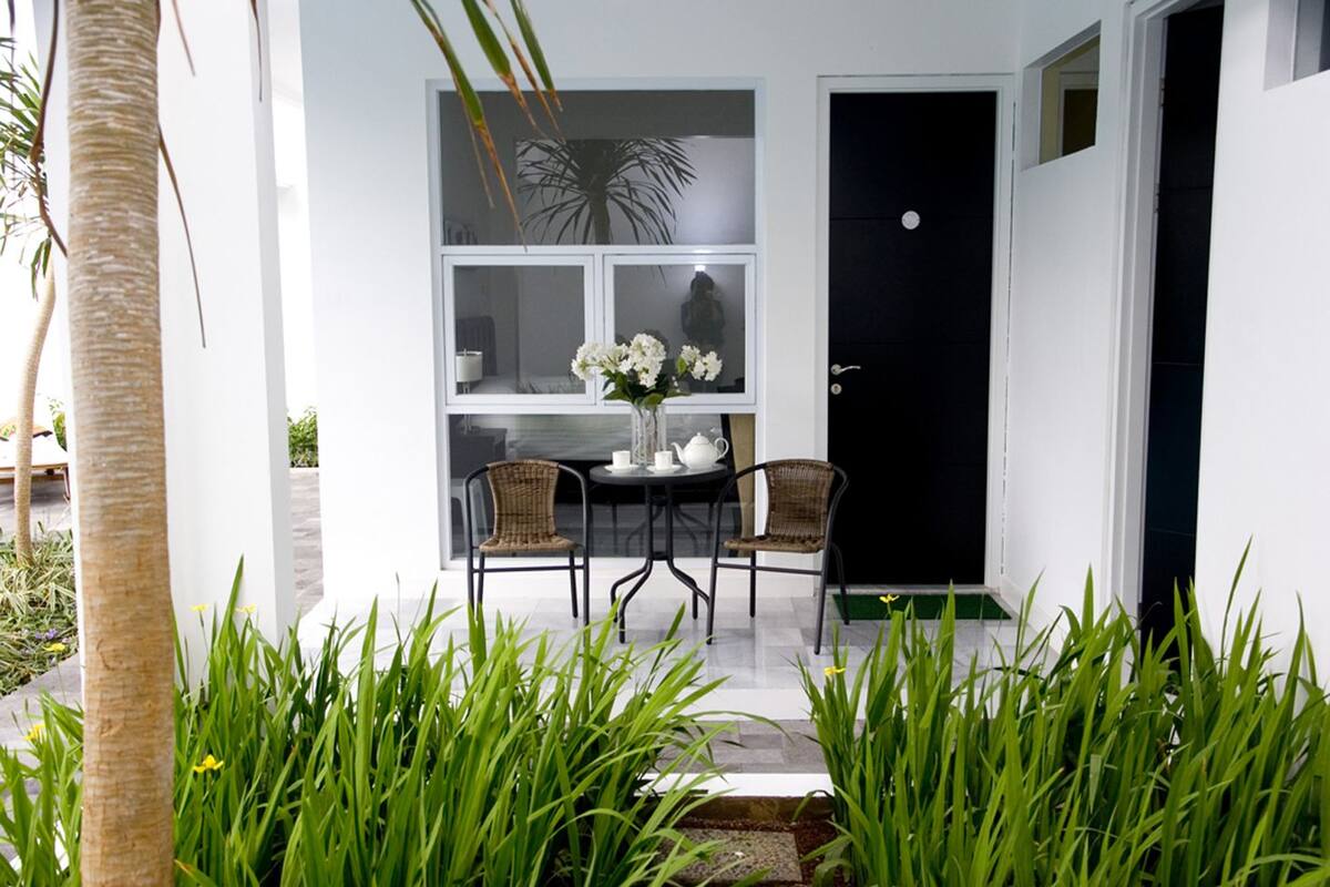 A modern outdoor seating area with a table and two wicker chairs, a black door, and a white wall with a window.