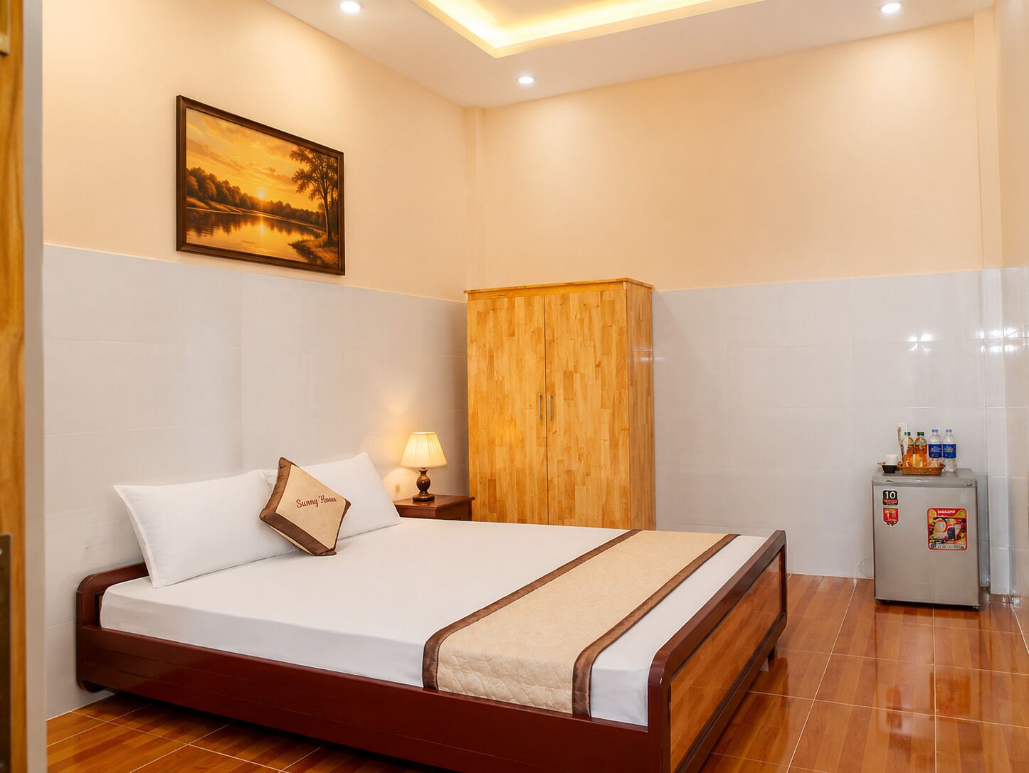 Une chambre d’hôtel avec un lit, une armoire en bois, une table de chevet avec une lampe, un réfrigérateur et un tableau encadré au mur.
