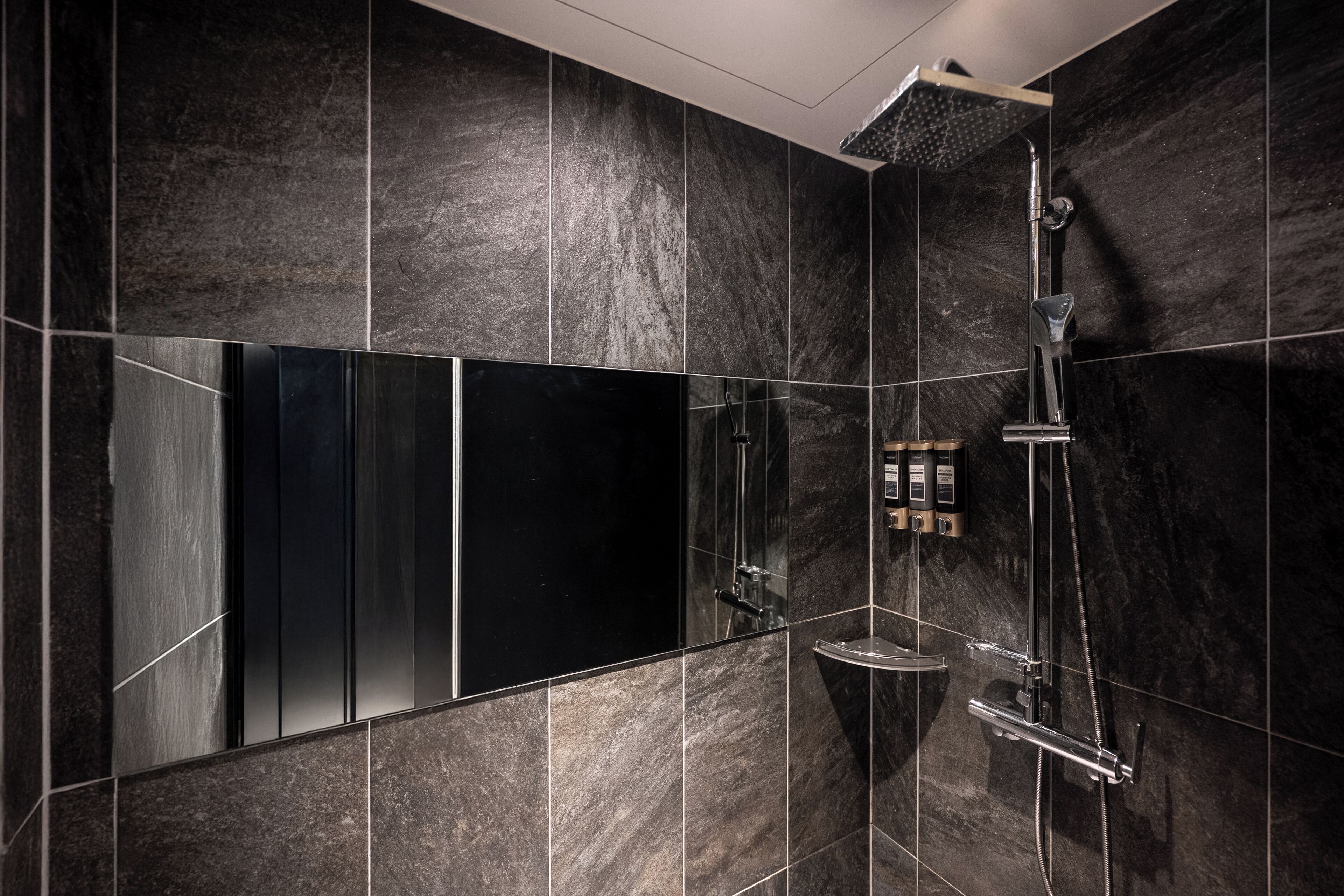 A modern bathroom with dark marble tiles, a large mirror, and a shower with a handheld showerhead.