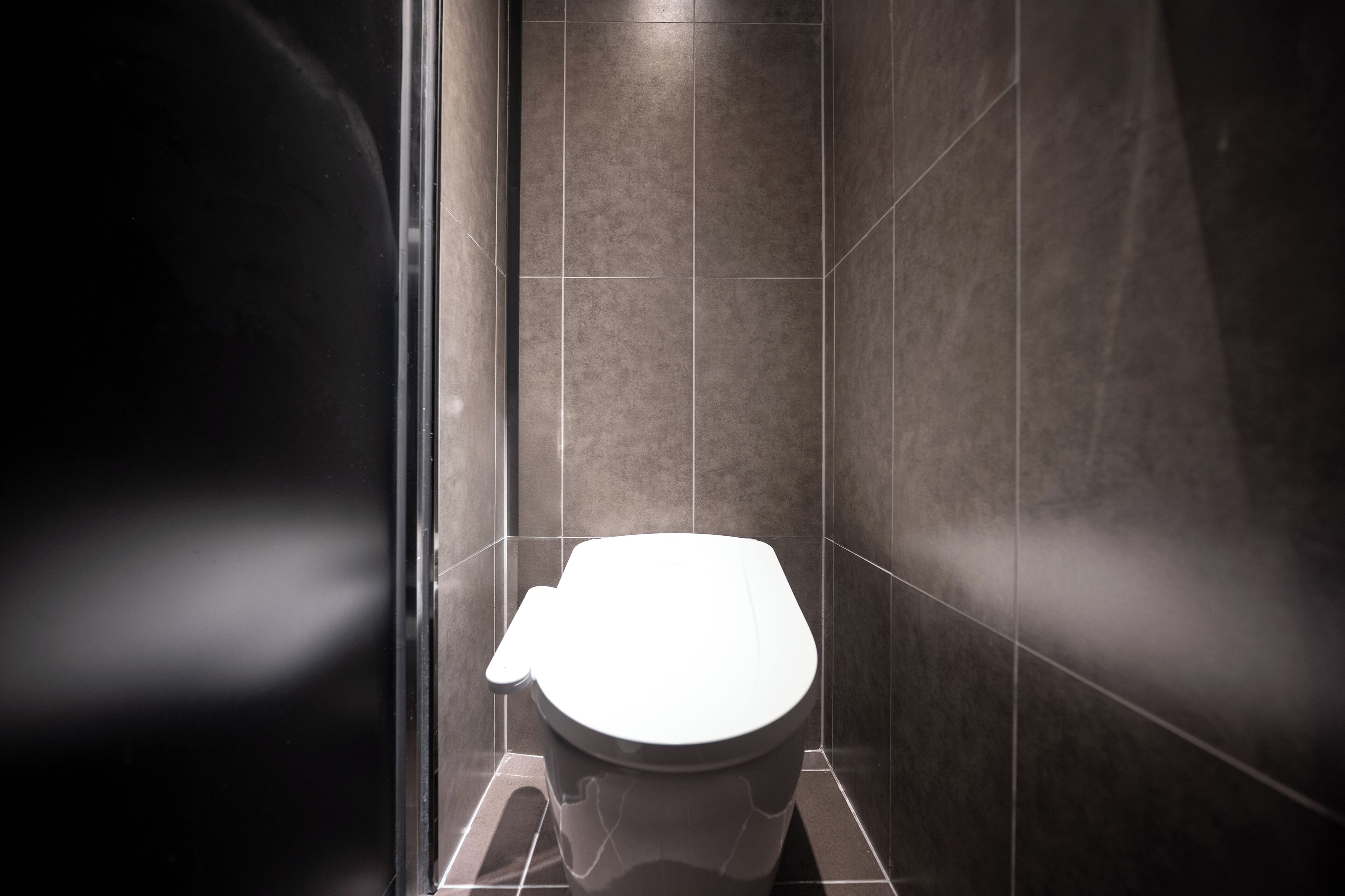 Un baño moderno con inodoro blanco, paredes de azulejos oscuros y cabina de ducha de cristal.