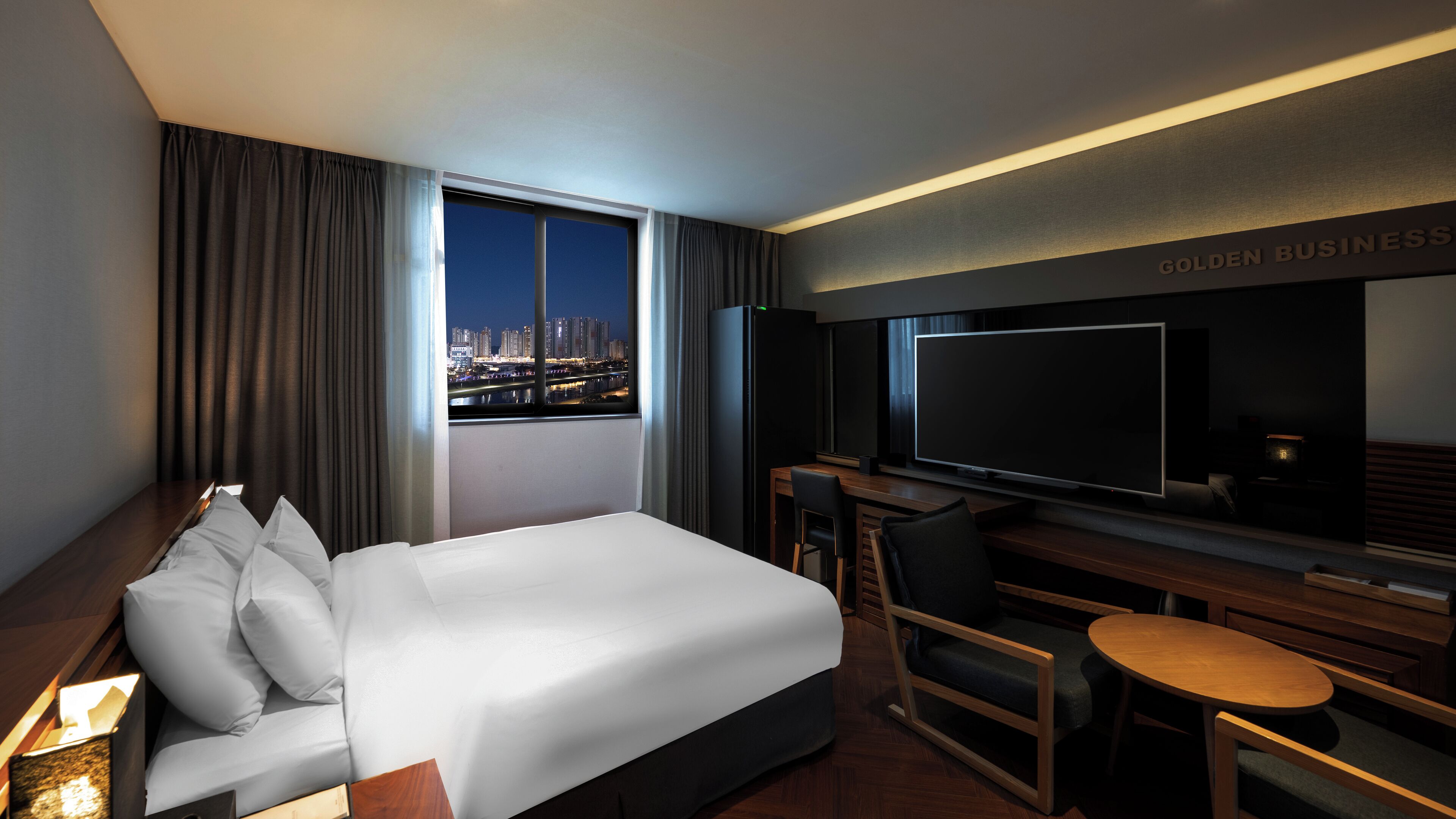 Una habitación de hotel con cama, escritorio con televisor y vistas a la ciudad. Un letrero dice 'GOLDEN BUSINESS'.