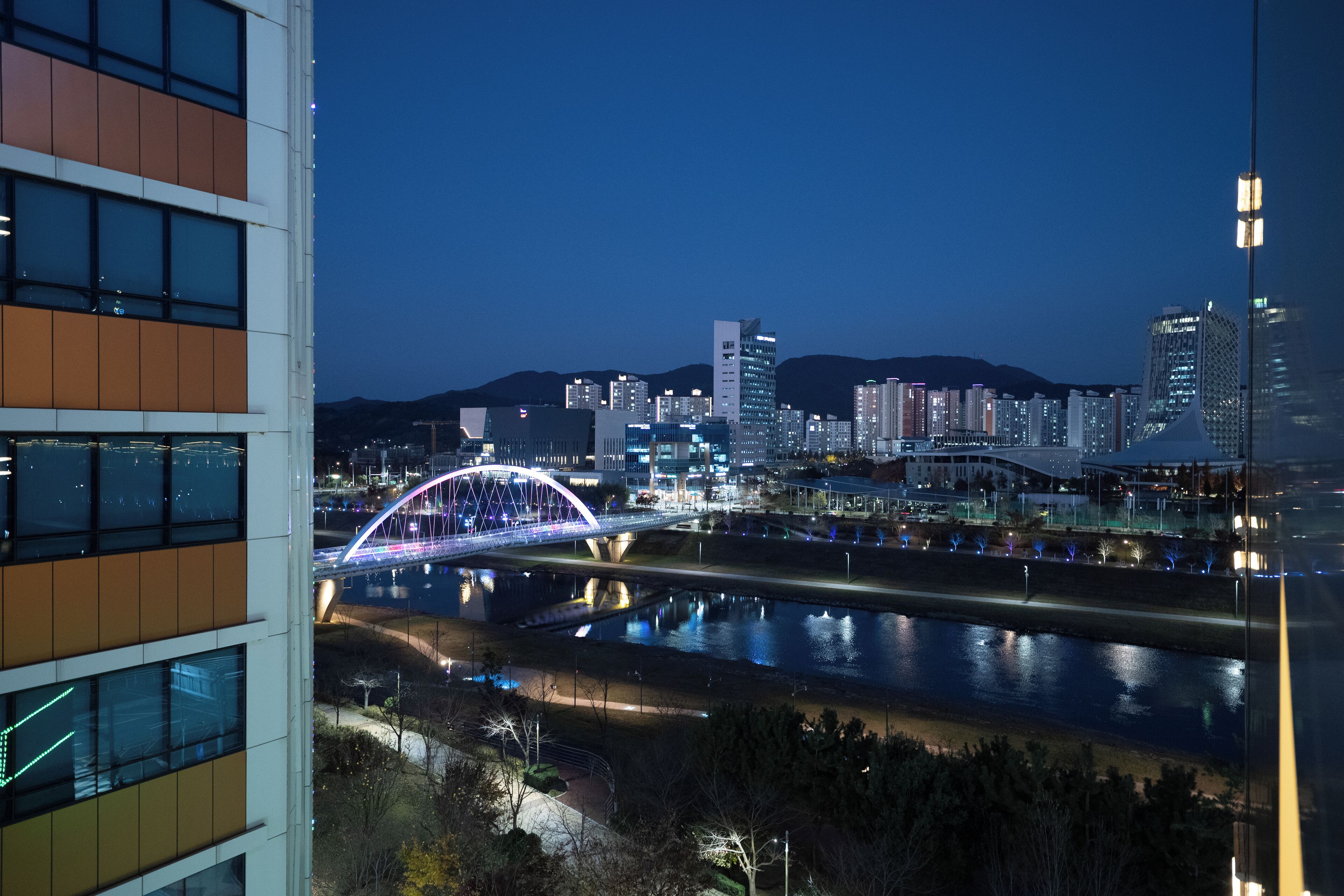 Paisatge urbà nocturn amb un pont brillant sobre un riu, edificis moderns i una vista clara de l'horitzó de la ciutat.