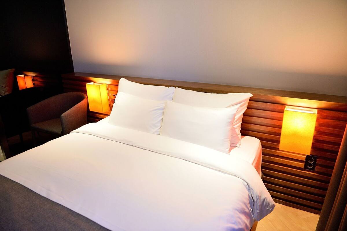 A neatly made bed with white linens, a wooden headboard, and two wall-mounted lamps.