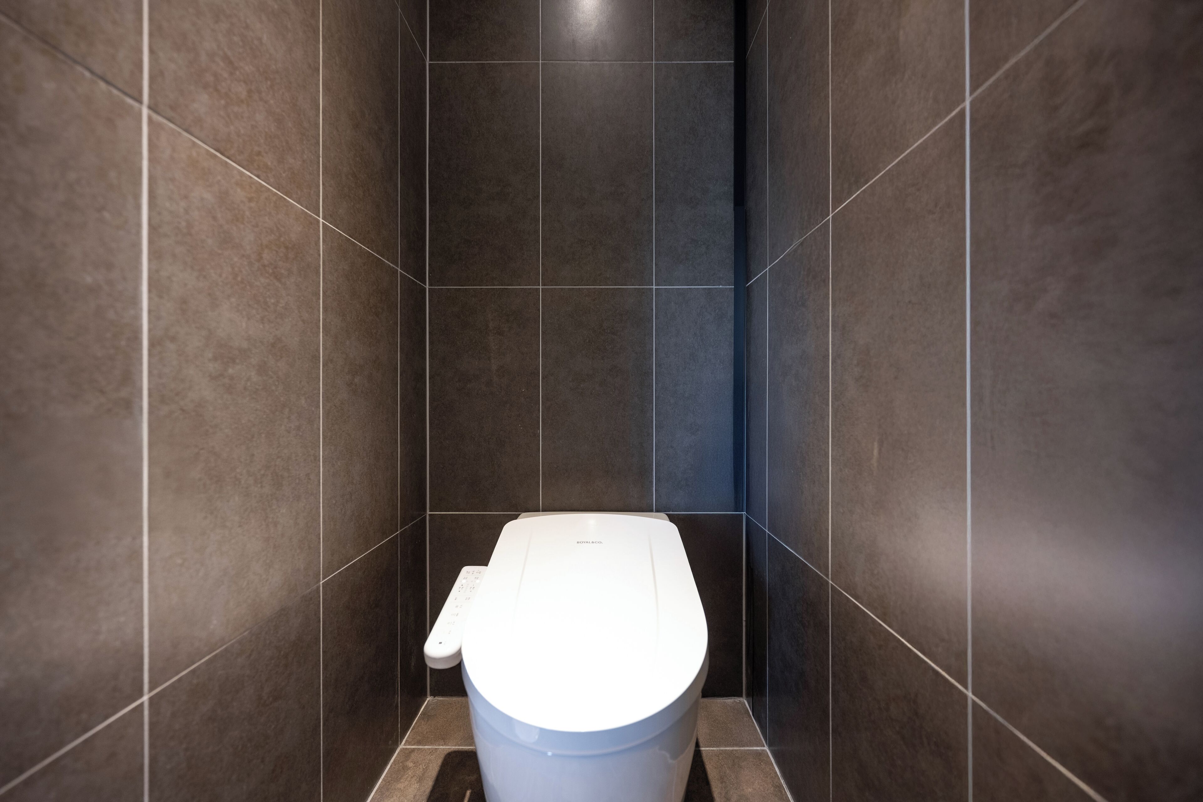 Un baño moderno con inodoro blanco, paredes de baldosas oscuras y un panel de control visible a la izquierda.