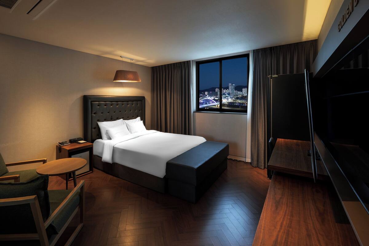 A hotel room with a large bed, a desk, a chair, a TV, and a view of the city at night.