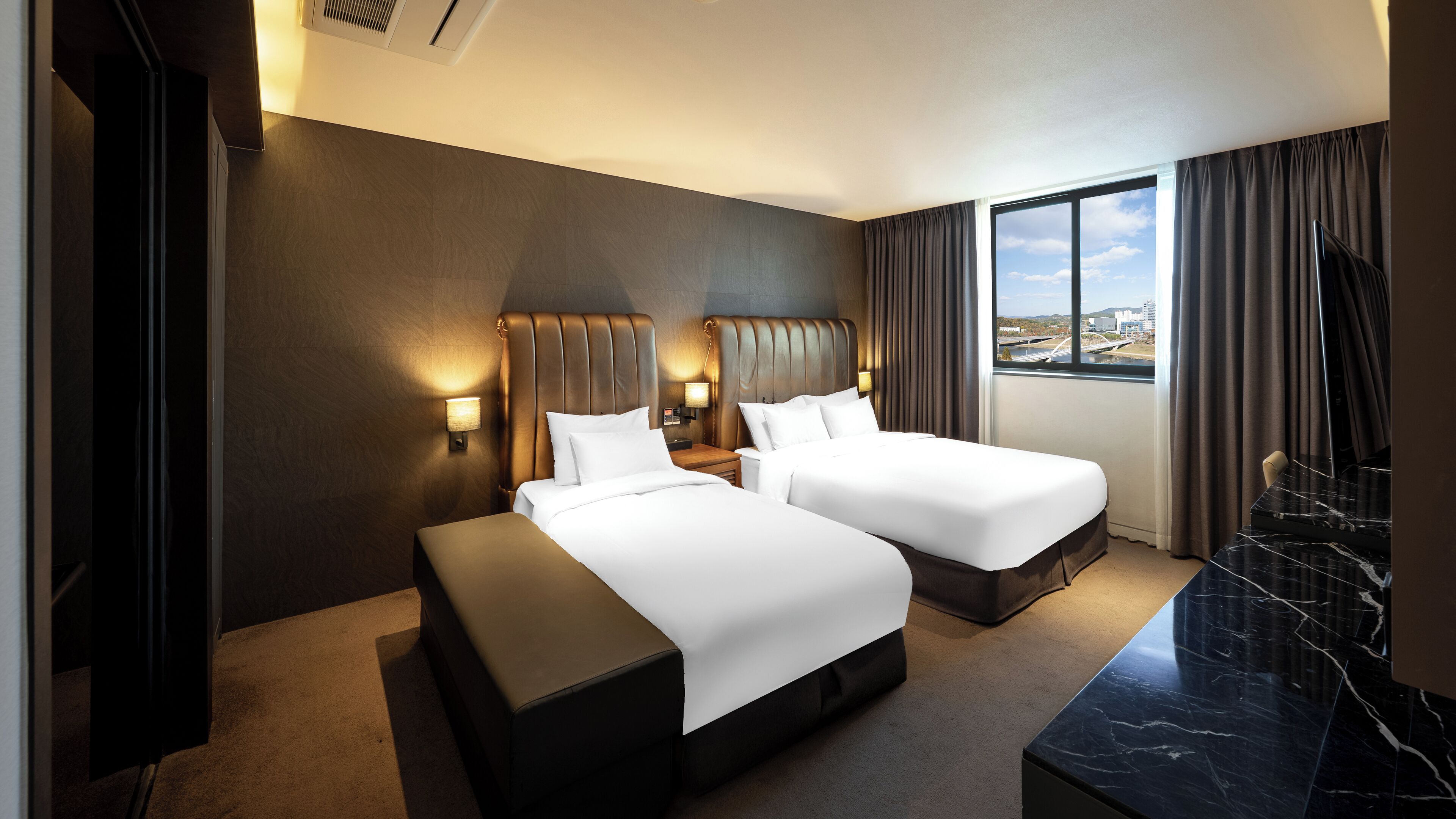 A hotel room with two beds, a large window with a city view, a marble side table, and a wall-mounted TV.