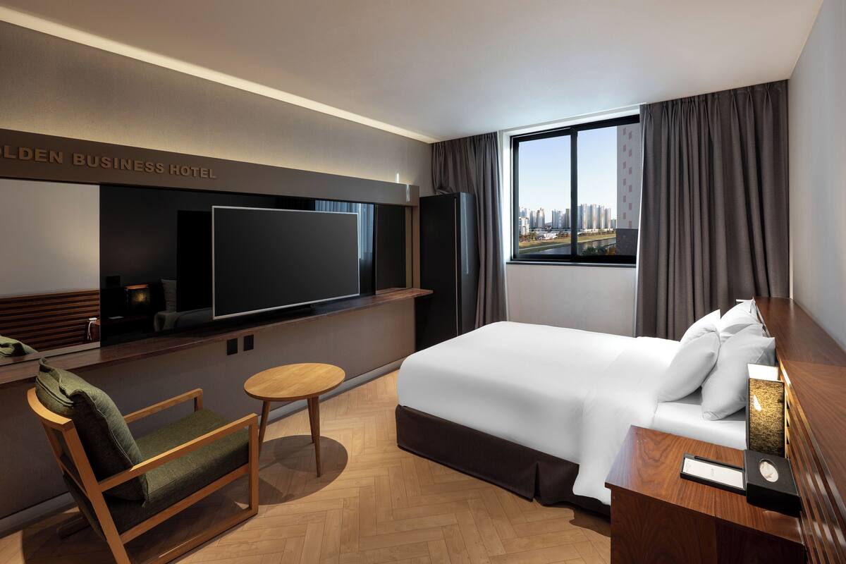 A hotel room with a bed, a TV, a chair, a small table, and a view of the city.