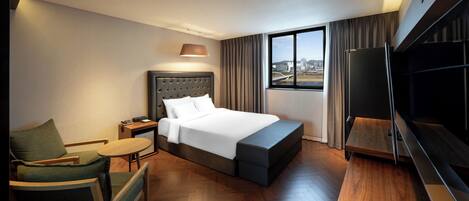 A hotel room with a bed, a desk, a chair, a TV, and a large window with a city view.