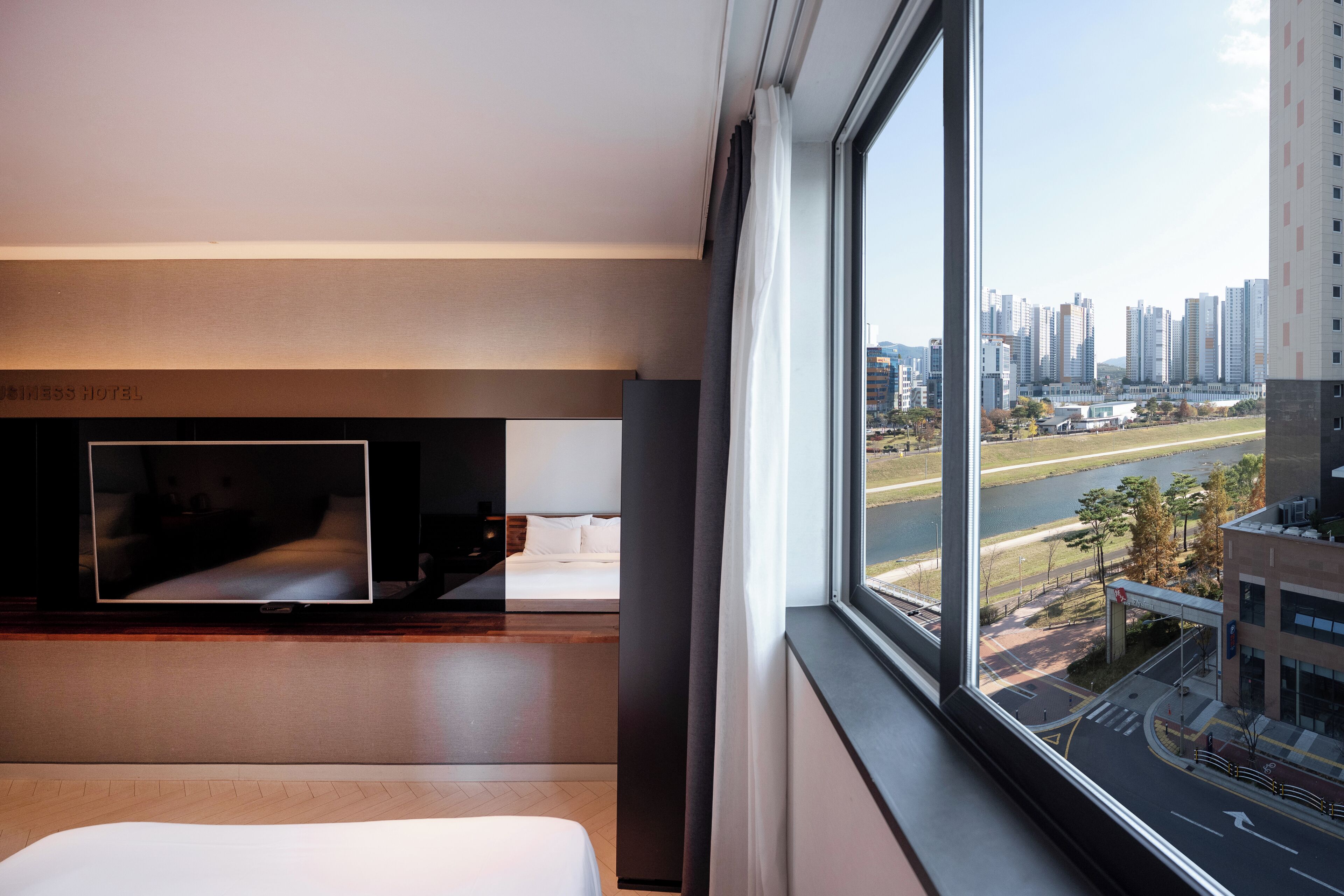 A hotel room with a bed, a TV, and a large window offering a view of a cityscape and a river.