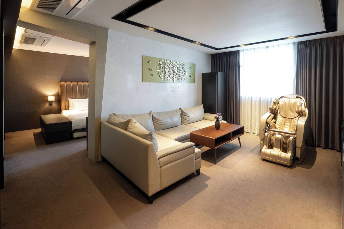 A modern hotel room with a sofa, a massage chair, a coffee table, and a bed.