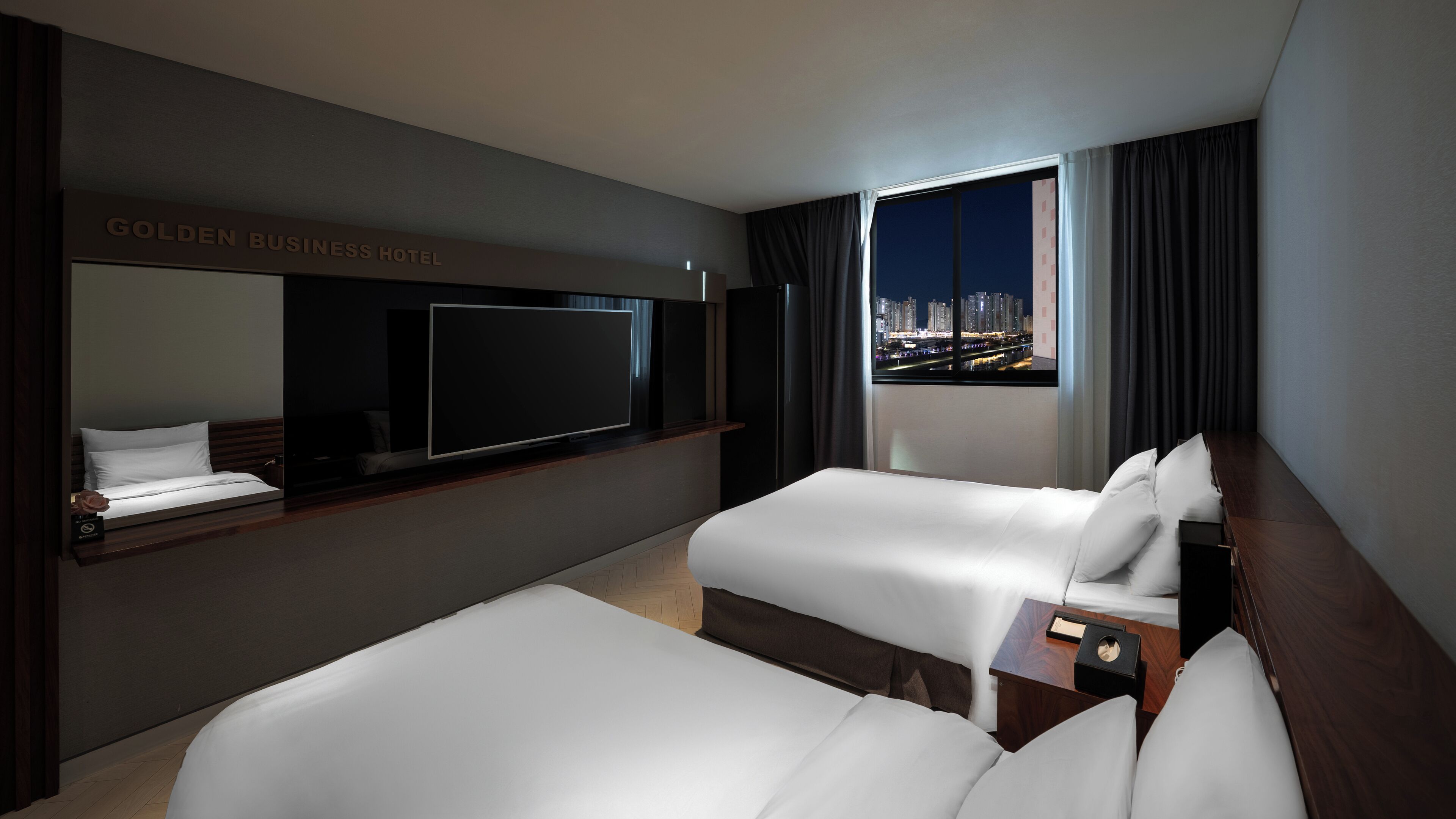 Quarto de hotel com duas camas, televisão de tela plana, uma janela grande com vista para a cidade e um letreiro com a inscrição "GOLDEN BUSINESS HOTEL".